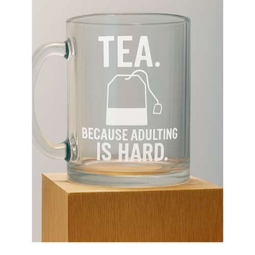 A clear glass mug with white text: 'TEA. Because adulting is hard.' sits on a wooden block.